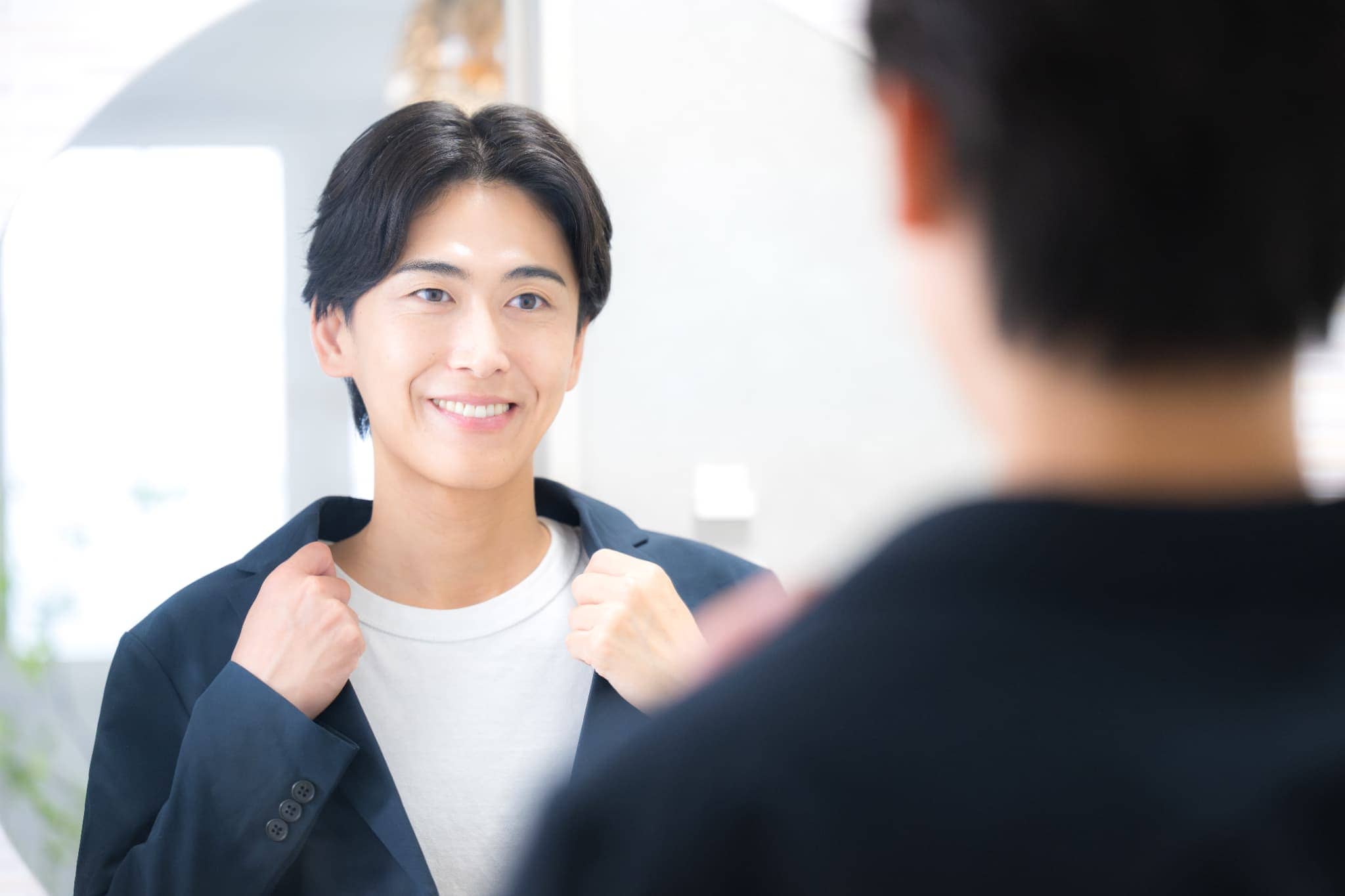 鏡の前でジャケットの襟元を整えながら、笑顔で自分の姿を見つめる若い男性